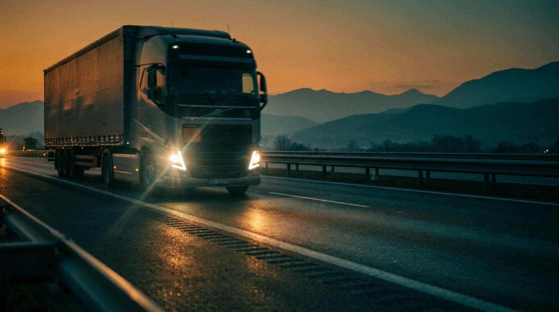A semi-truck at speed on a dusk highway, headlights flaring on wet asphalt